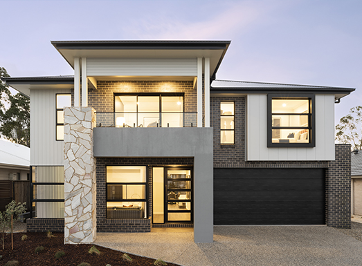 Modern two-story residential house with white and dark brick facade, large black-framed windows, a prominent stone accent column, black garage door, and illuminated interior spaces at dusk.