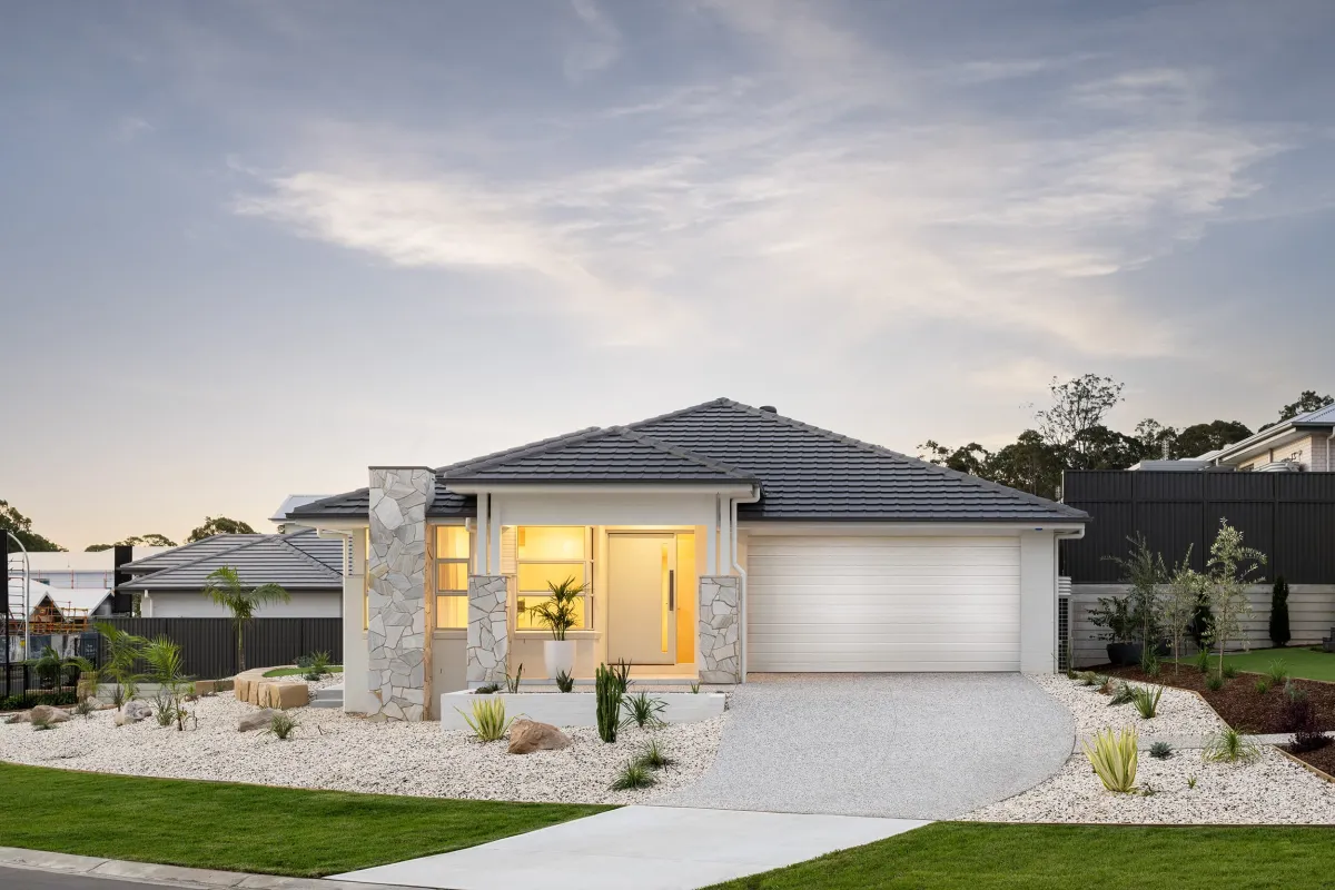 Modern single-story display home with white rendered walls and dark blue-grey tile roof, featuring stone accent pillars, large illuminated windows, and a double garage. The front yard includes a gravel landscaped garden with native plants, succulents, and decorative rocks, with green lawn on either side. Neighboring homes visible in the background under a partly cloudy sky.