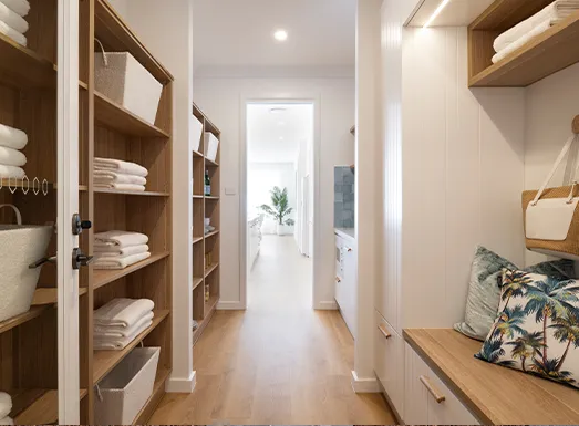 Modern mudroom with built-in wooden shelving on left side storing folded linens and towels, wooden bench with storage on right side, hardwood flooring, recessed lighting, and view through doorway to bright entryway with potted plant.