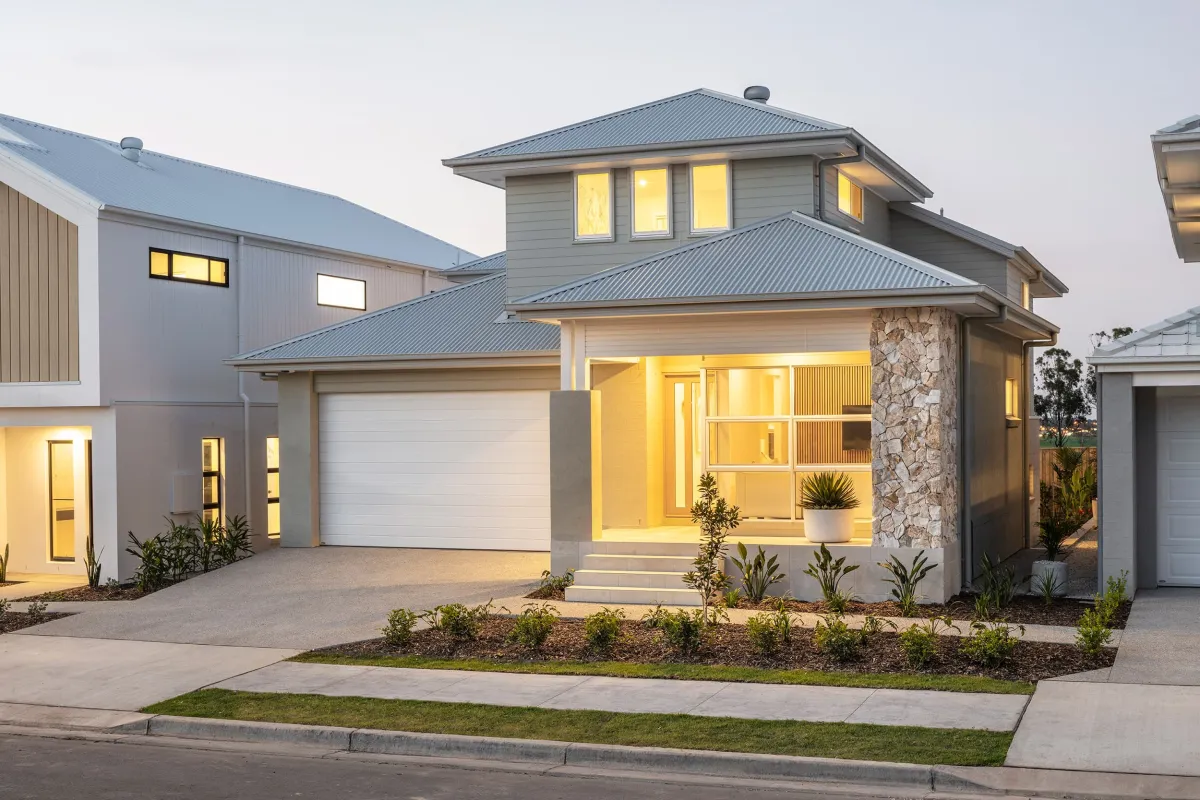 Modern two-story residential home at dusk with illuminated interior lights. The house features a gray and white exterior with metal roof, stone accent wall, white garage door, and large windows. Landscaped front yard with young plants and shrub borders. Adjacent homes visible on either side of the street.