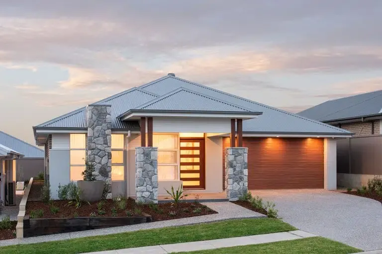 Modern single-story display home with white exterior walls, blue metal roof, and stone pillars flanking the illuminated entrance door. Features a brown garage door on the right, landscaped front garden with native plants, and gravel driveway at dusk.