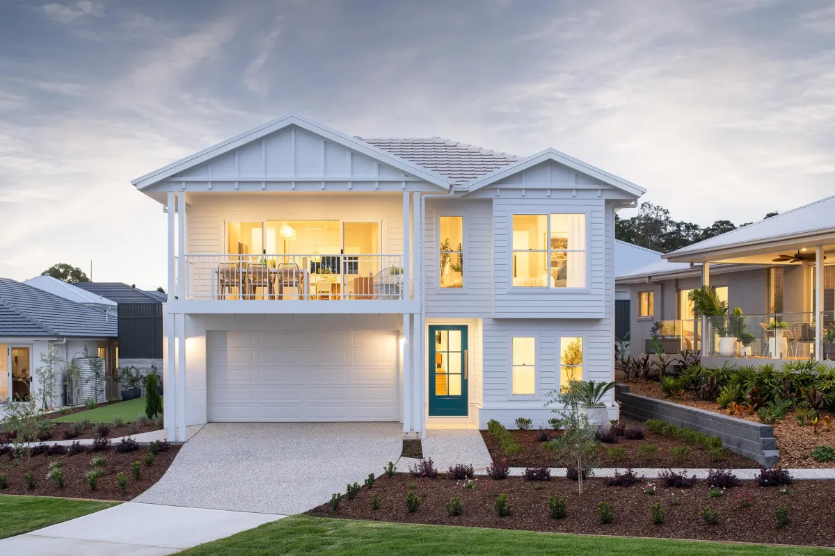 Front facade of the Ravenna 264 display home in Warnervale, a modern two-story white weatherboard house with pitched roofs, featuring a double garage, upper-level balcony with metal railing, illuminated windows, dark green front door, and manicured landscaping with young plants and mulch.