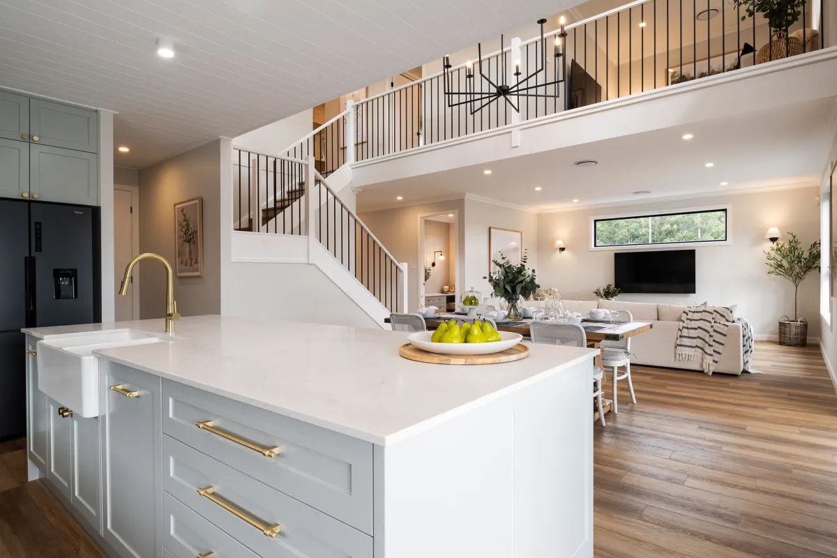 Modern open-plan kitchen and living area in a display home featuring a white kitchen island with gold fixtures, gray cabinetry, black appliances, and white countertops. The space flows into a living room with a TV, comfortable seating, and large windows. A staircase with black metal railings leads to an upper level mezzanine. Wooden flooring throughout, warm lighting, and fresh green plants complete the contemporary design.