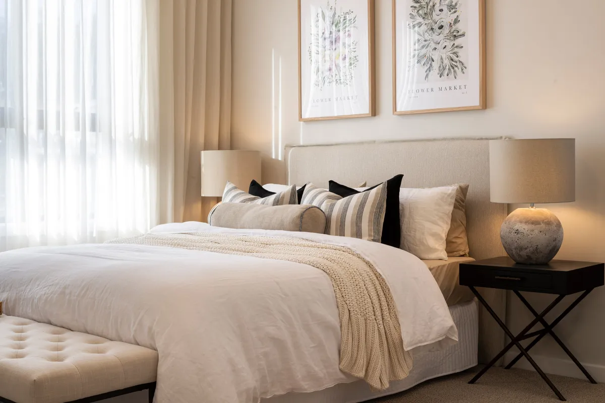 Master bedroom in Montgomery Homes Waverley 324 display home featuring a neutral upholstered bed with striped and black pillows, white bedding with beige throw blanket, matching nightstands with spherical lamps, botanical artwork above headboard, floor-to-ceiling curtains, and modern minimalist décor in warm neutral tones.