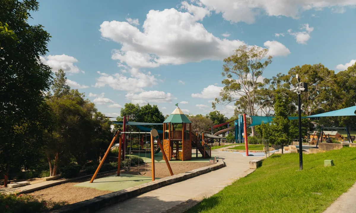 Millfield Rise Estate playground featuring a large multi-level play structure with orange and red wooden components, green roof tower, and slide, surrounded by trees, grass areas, paved pathways, and blue shade sails on a sunny day with blue sky and white clouds.