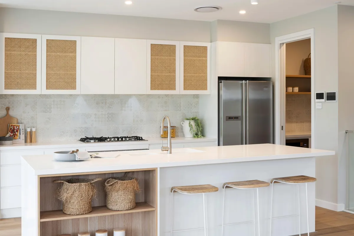 Modern luxury kitchen with white cabinetry featuring rattan-front upper cabinets, light marble backsplash, stainless steel refrigerator, gas cooktop, and a large white quartz island with three wooden bar stools. Open shelving displays woven baskets and decorative items. Warm wood accents and recessed lighting complete the contemporary resort-style design.