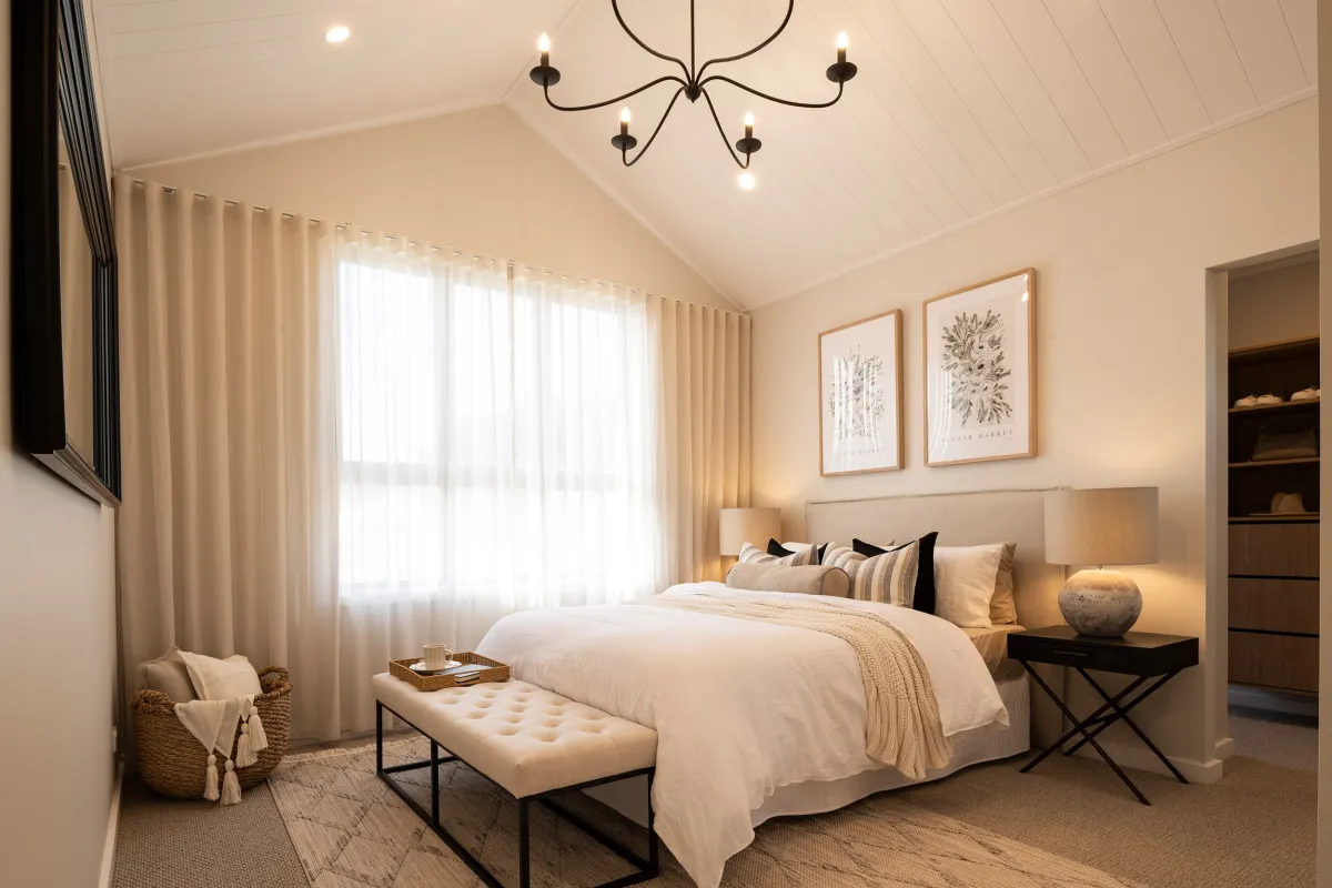 Montgomery Homes Waverley 324 display home master bedroom featuring a bed with neutral bedding, upholstered headboard, decorative pillows, and a bench at the foot. The room has cream-colored walls, vaulted ceiling with black chandelier, large windows with cream curtains, framed botanical artwork, bedside tables with lamps, and built-in wooden cabinetry.