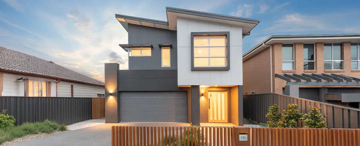 Modern two-story house in Adamstown with contemporary design featuring white upper level and dark gray lower level, illuminated entry door, garage, metal fencing, and neighboring brick homes visible on either side at dusk.