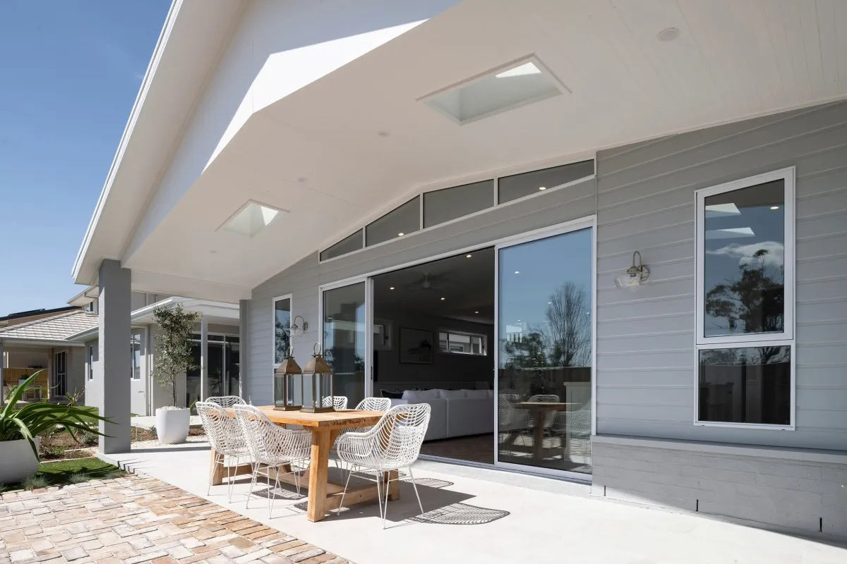 Modern display home exterior featuring a covered alfresco dining area with wooden table and white woven chairs, large sliding glass doors, grey cladding, white ceiling with skylights, brick paving, and landscaping at Oran Park.
