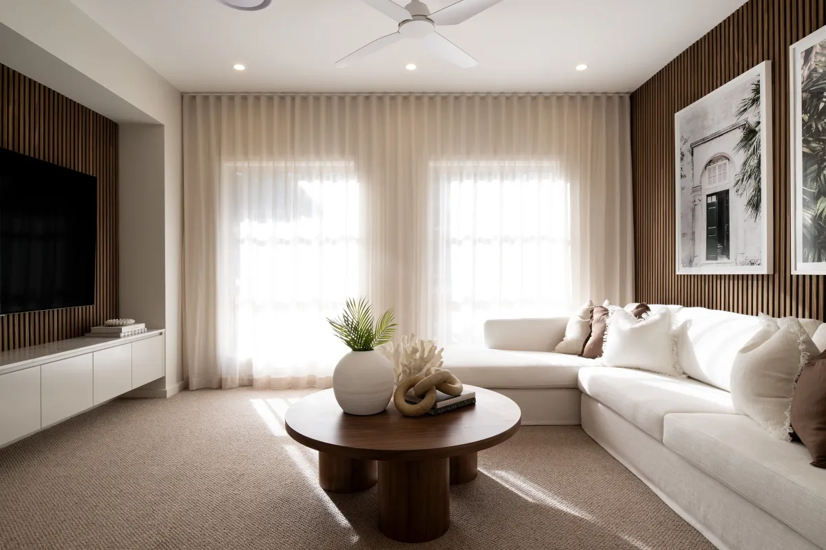 Modern living room with cream-colored sectional sofa, round dark wood coffee table with decorative plant and sculpture, white media console with flat-screen TV on left wall, vertical wood slat accent walls, sheer cream curtains over large windows, recessed ceiling lights, ceiling fan, framed botanical artwork on right wall, and neutral carpeting.