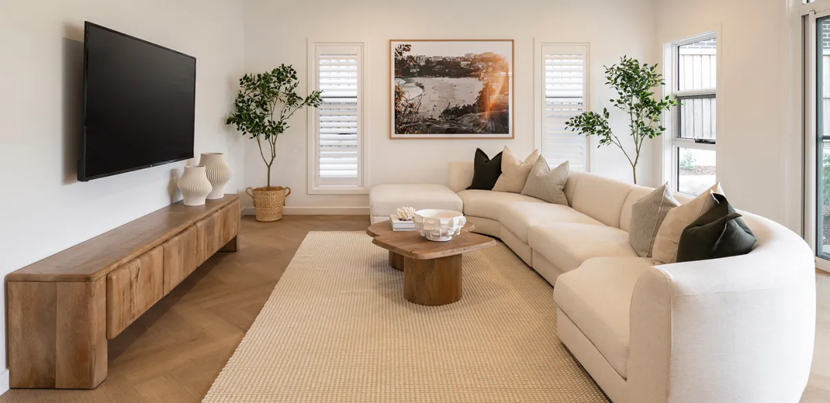 Modern lounge room with curved cream sectional sofa, wooden media console with mounted flat-screen TV, round wooden coffee table on neutral area rug, framed landscape artwork, potted plants, white shuttered windows, and natural wood flooring.