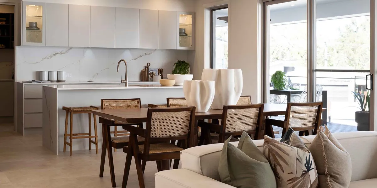 Modern open-plan kitchen and dining area in Ridgeview 321 display home featuring white cabinetry with marble backsplash, bronze faucet, dining table with rattan-backed chairs, cream upholstered sofa with patterned pillows in foreground, and large sliding glass doors opening to outdoor balcony with landscape views.
