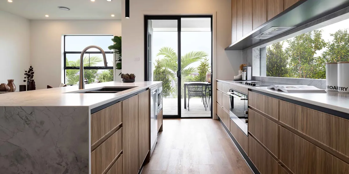 Modern kitchen with light wood cabinetry, white marble countertops, and stainless steel appliances. A central island with sink and curved faucet anchors the space. Large windows and glass doors overlook a tropical garden with palm trees and outdoor seating area. White walls, recessed lighting, and a mirrored backsplash complete the bright, contemporary design.