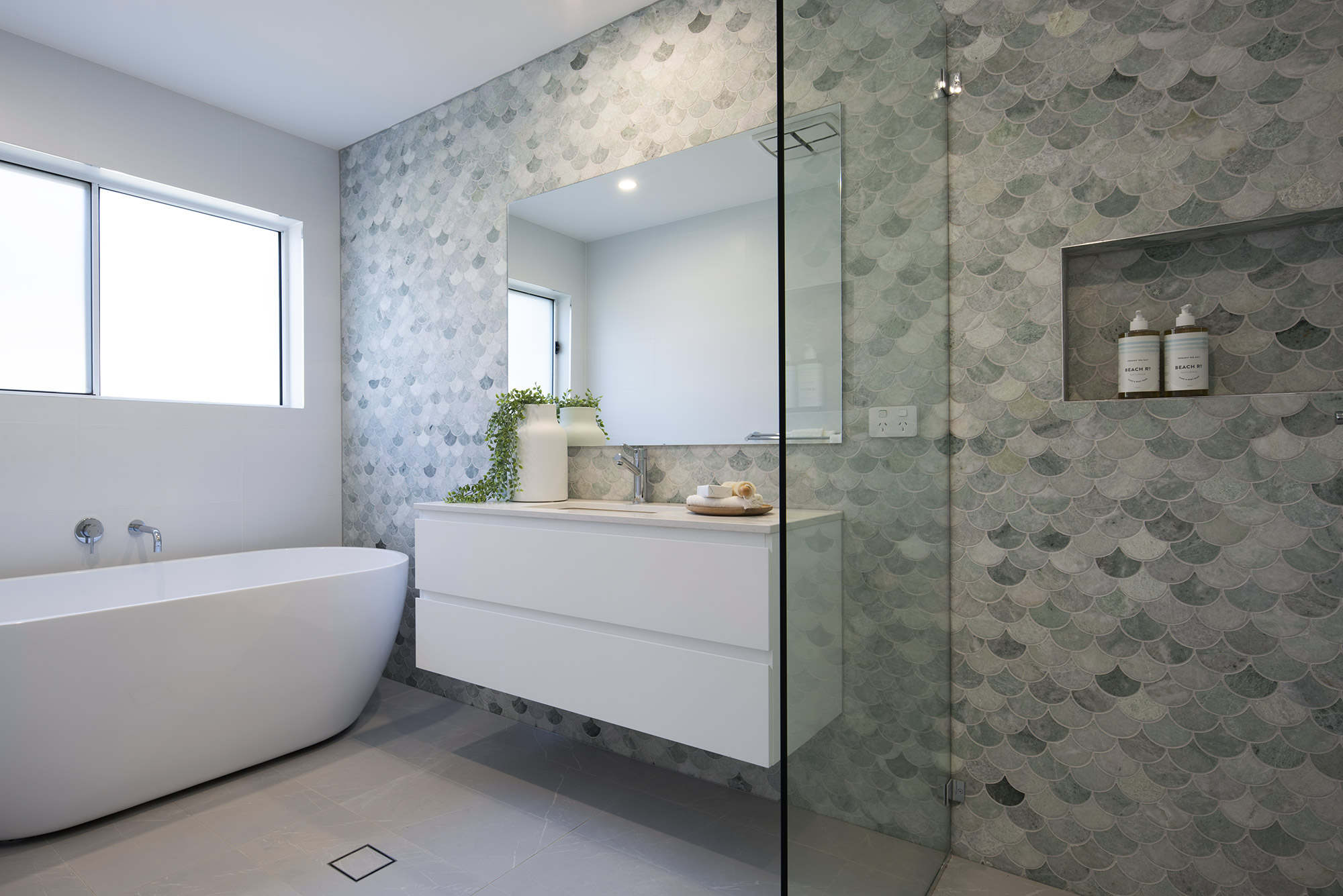 Modern spa-style bathroom featuring a white freestanding soaking tub on the left, a white floating vanity with large mirror in the center, and a glass-enclosed shower with seafoam green scallop-patterned tile on the right. Natural light enters from a window above the tub. The walls are covered in matching scallop-patterned wallpaper in soft green and gray tones. A potted plant and decorative items sit on the