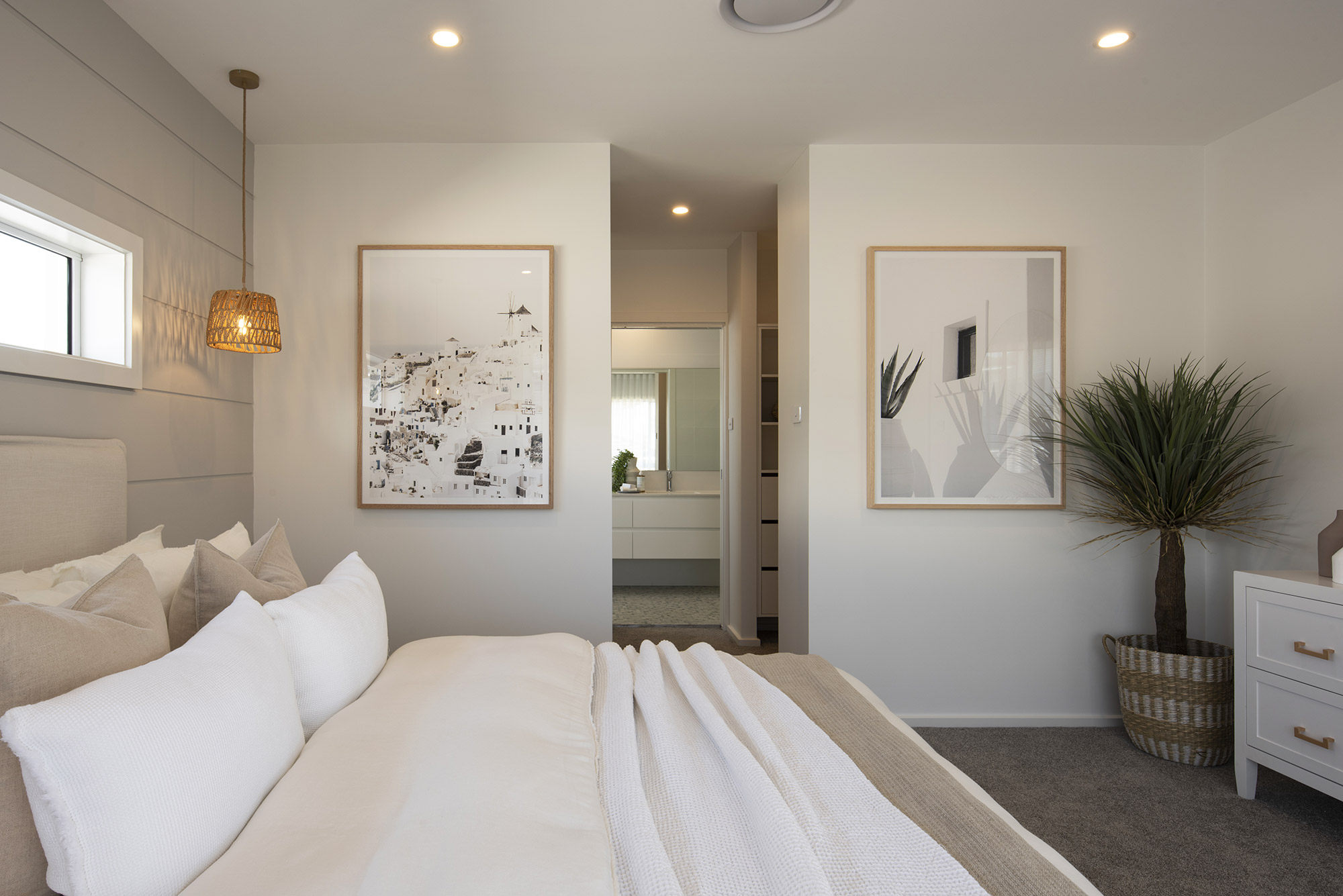 Modern bedroom with neutral tones featuring a bed with white pillows and cream bedding, woven pendant light, framed coastal artwork on walls, ensuite bathroom visible through open doorway, potted plant with dark foliage in woven basket, white dresser, and recessed ceiling lights.