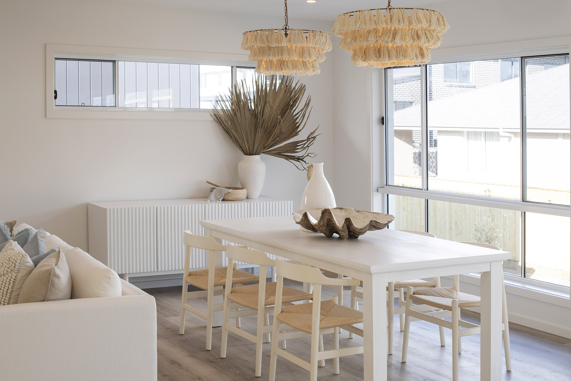Bright coastal dining room with white rectangular table and natural wood chairs, cream-colored sofa with pillows on left, two layered raffia pendant lights overhead, dried palm fronds in white vase on radiator shelf, decorative shell bowl on table, and large glass sliding doors opening to outdoor deck with ocean views.