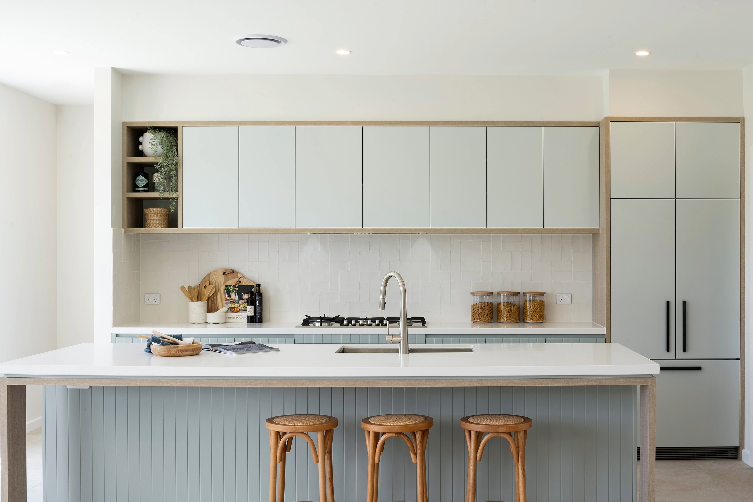 Modern coastal kitchen with light blue and white cabinetry, white countertops, and a gas cooktop. Open shelving displays decorative items and kitchen essentials. Three wooden bar stools sit at a white island. Recessed lighting and clean lines create a bright, minimalist design.