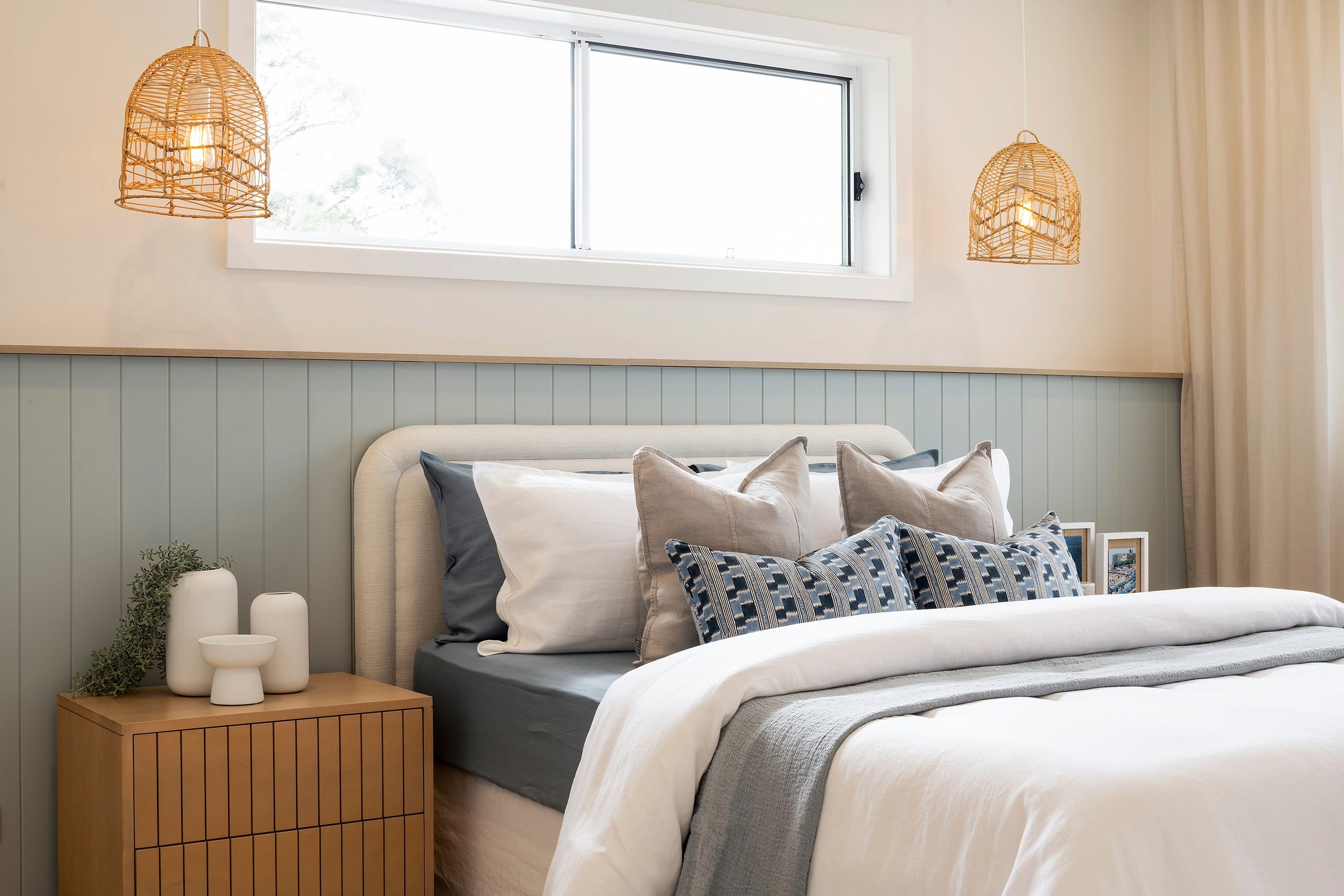 Coastal-style bedroom with a curved upholstered headboard, layered bedding in neutral and blue tones, wooden nightstand with white ceramic decor, sage green shiplap wall paneling, two woven rattan pendant lights flanking a large horizontal window, and cream-colored walls.