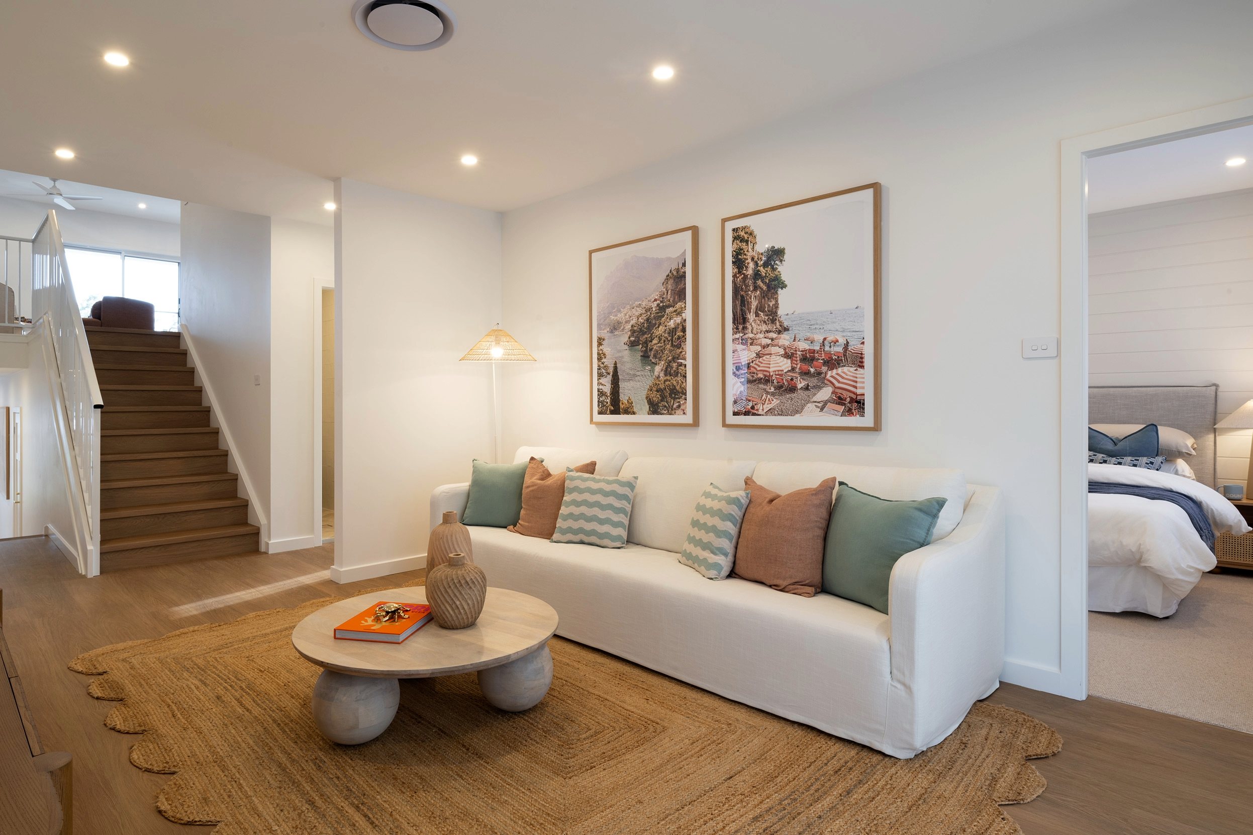 Modern living room with cream-colored sofa adorned with green and orange pillows, facing a round wooden coffee table on a woven rug. Two framed coastal landscape photographs hang on the white wall above. A wooden staircase with white railing is visible on the left, and a bedroom with a made bed can be seen through an open doorway on the right. Recessed ceiling lights and a pendant lamp illuminate the bright, open-plan space.
