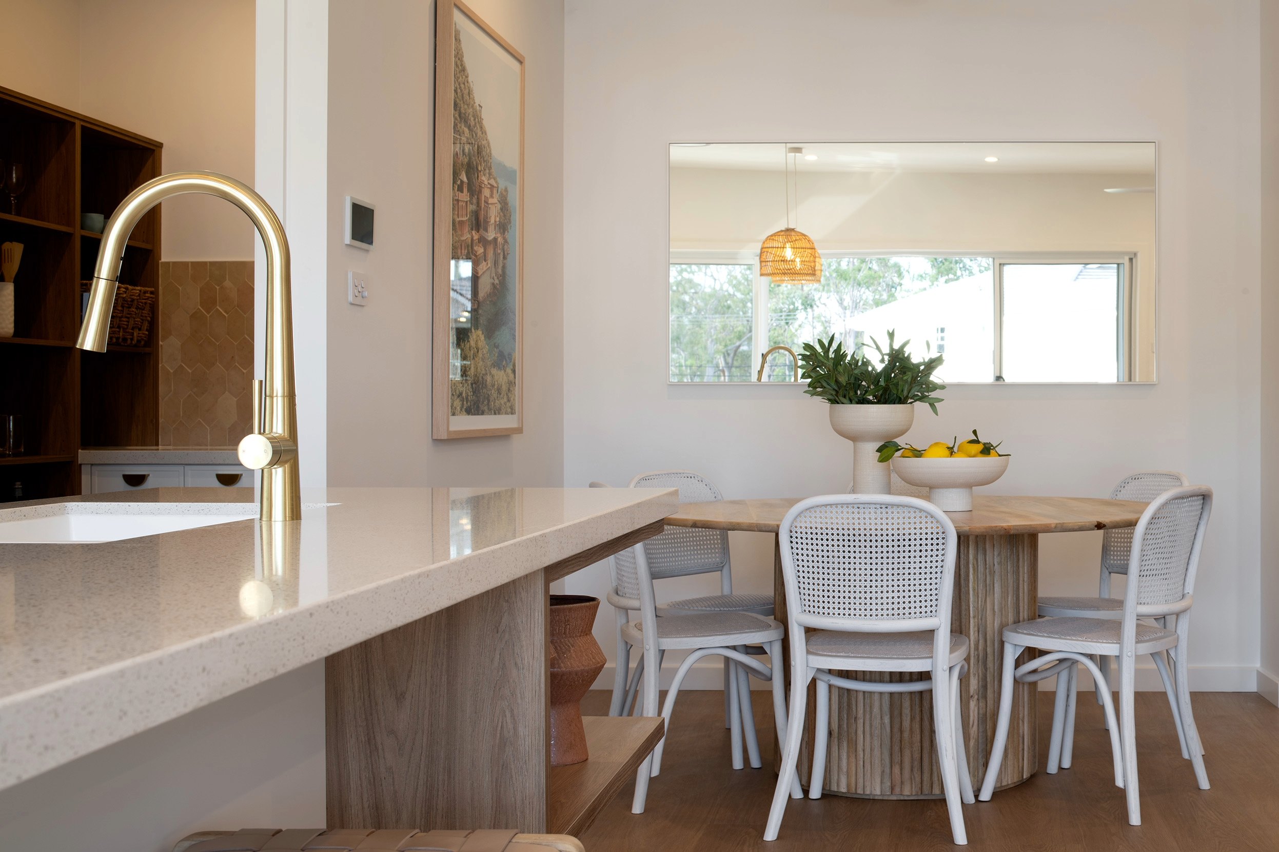Modern open-plan kitchen and dining area with light wood dining table, white rattan chairs, cream stone countertop with brass pull-down faucet, pendant lighting, potted plants with lemons, framed artwork, and large windows overlooking landscaped grounds.