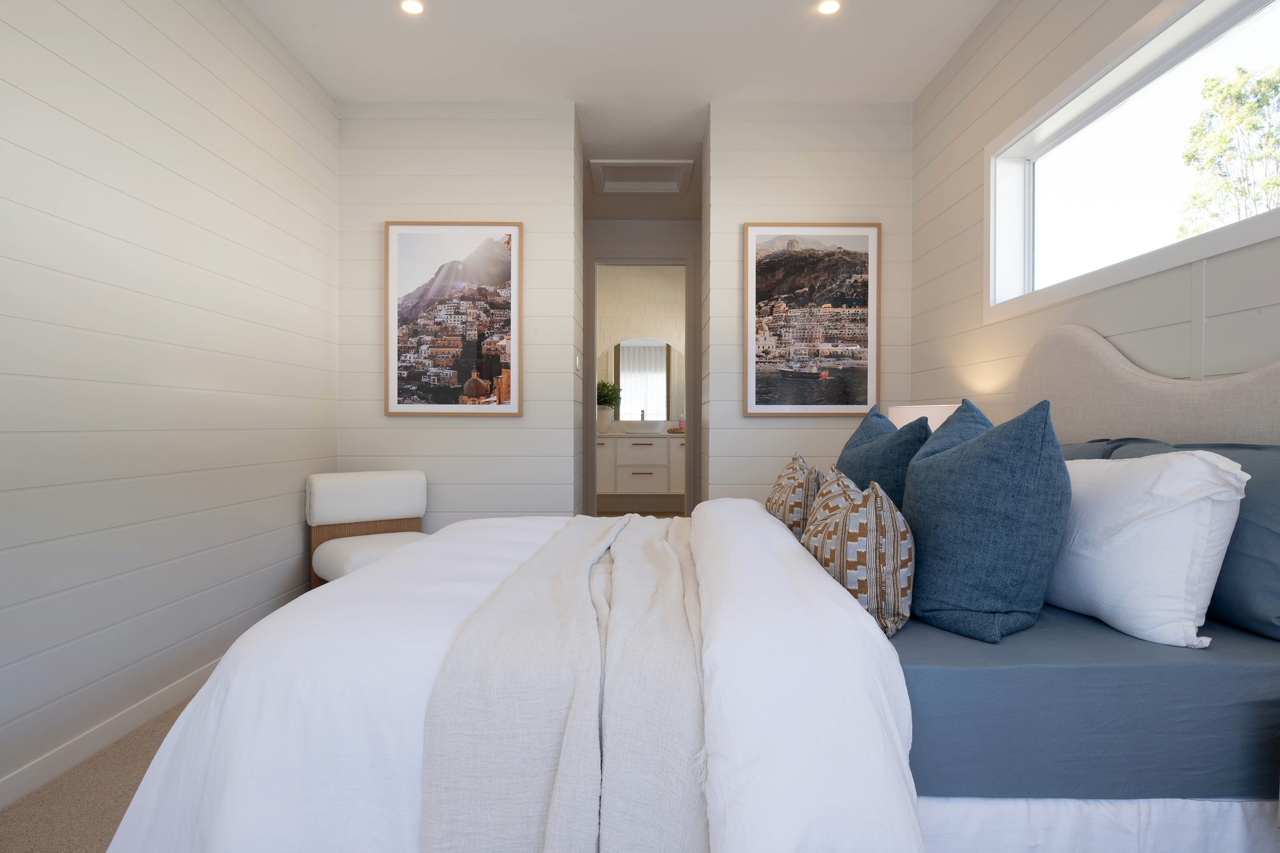 Modern bedroom interior with two beds featuring white bedding and blue pillows, white shiplap walls, framed coastal landscape photographs, a central ensuite doorway, recessed ceiling lights, and natural light from a side window.