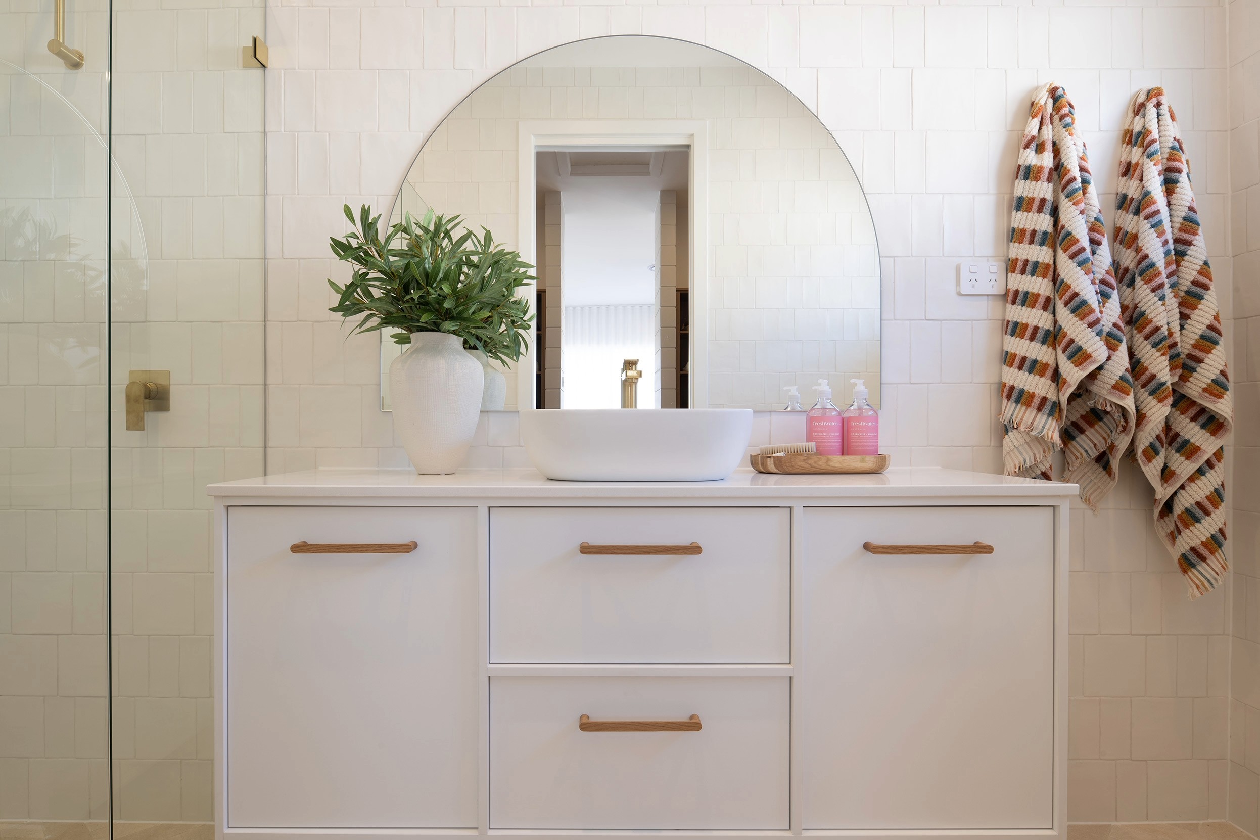 Modern coastal bathroom with white subway tile walls, white vanity with wooden handles, round mirror, white vessel sink, potted green plant, pink toiletries, patterned towels, and glass shower enclosure with brass fixtures.