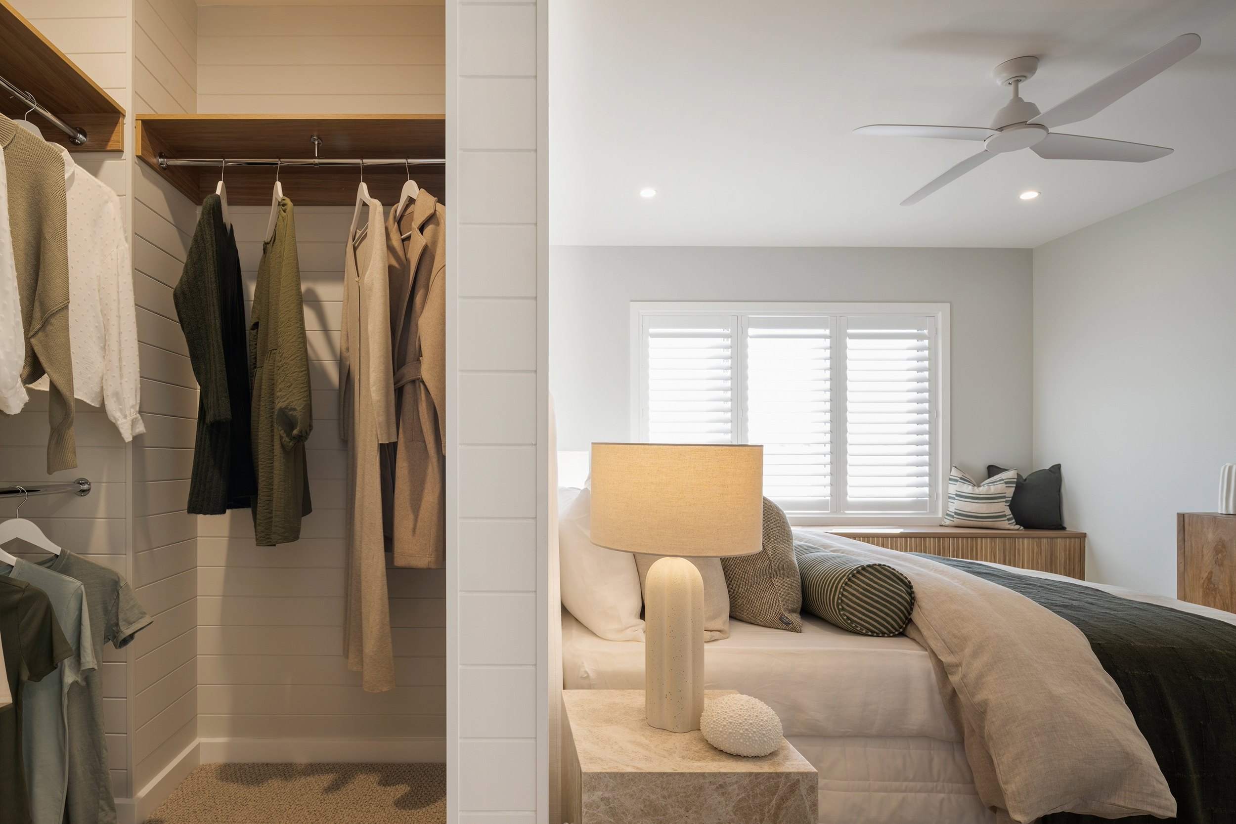 Luxurious modern bedroom with adjoining walk-in closet featuring white subway tile walls, wooden shelving with hanging neutral-toned clothing, and a bed with cream bedding, decorative pillows, and a table lamp with yellow shade. Large shuttered windows and ceiling fan visible in the bright, minimalist space.