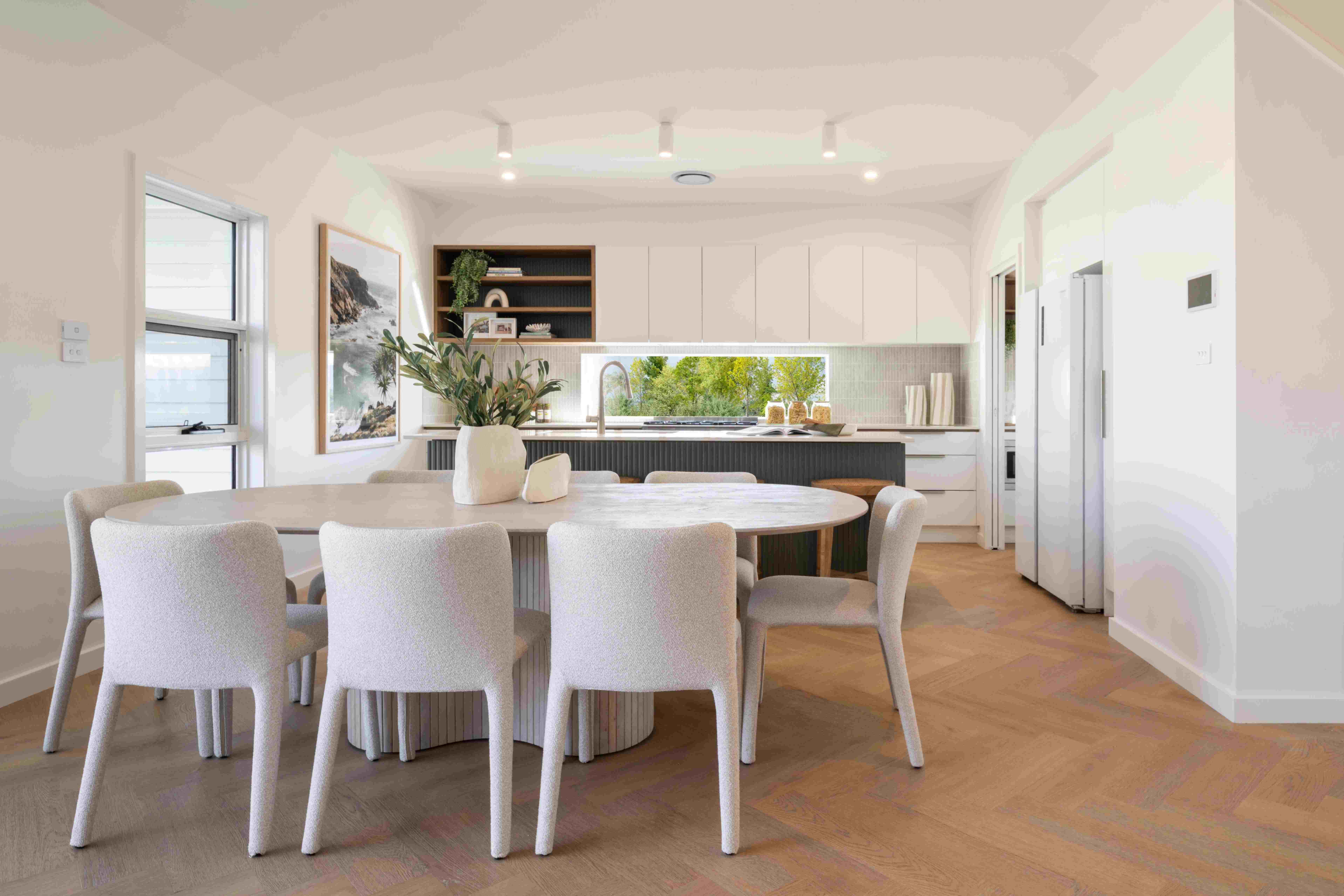 Modern kitchen and dining area with a round marble table surrounded by four cream-colored upholstered chairs on wooden flooring. White cabinetry lines the back wall with a large window overlooking greenery, open shelving with decor, and a cream ceramic vase centerpiece. Recessed ceiling lights illuminate the bright, minimalist space with coastal design elements.