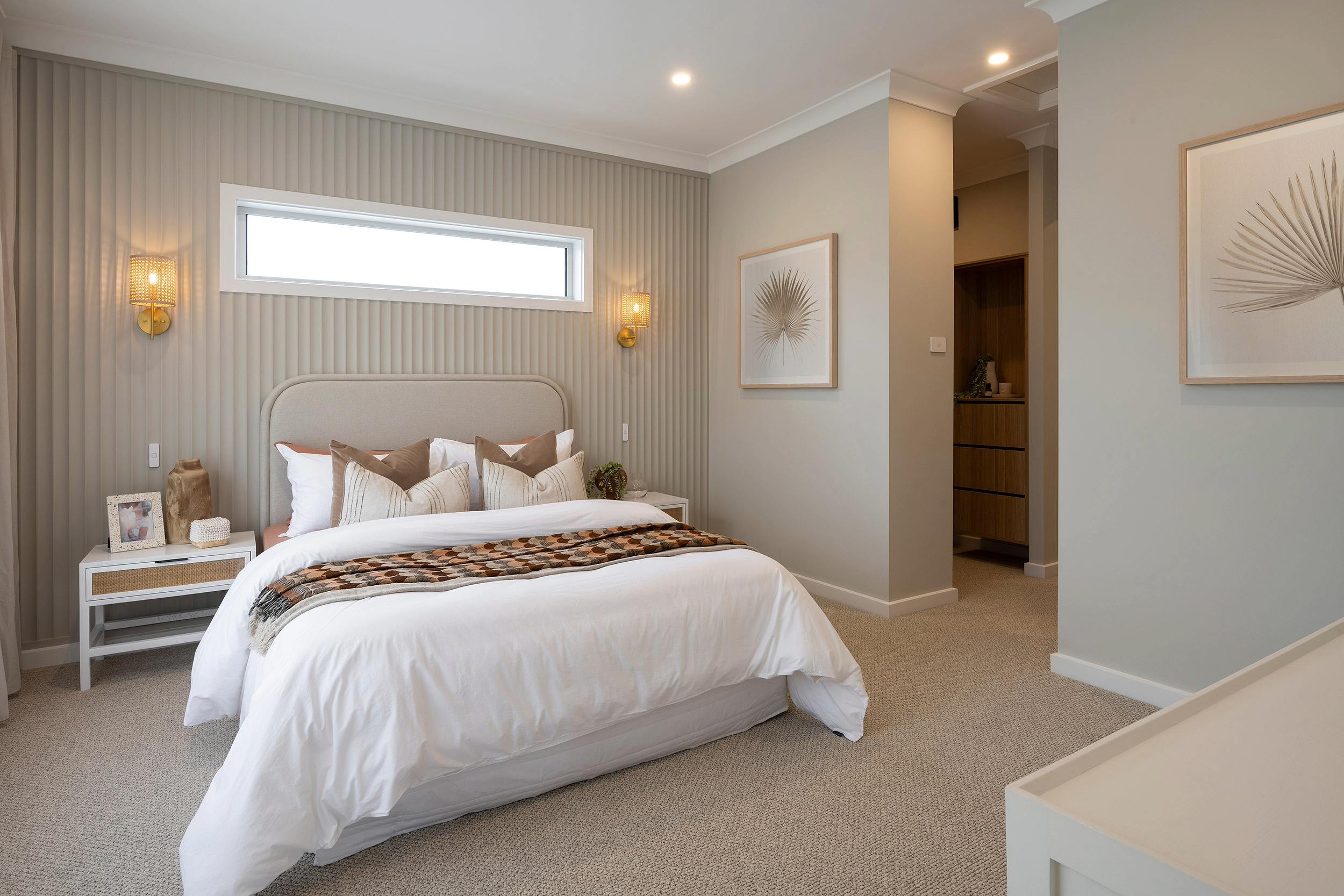 Luxurious resort bedroom with a queen bed featuring white bedding and decorative pillows, upholstered headboard, and a woven throw blanket. The room includes a white nightstand with decor, gold wall sconces, vertical striped wallpaper, and framed botanical artwork. A horizontal window provides natural light, and an ensuite bathroom entrance is visible on the right. The space features warm neutral tones, recessed lighting, and beige carpet throughout