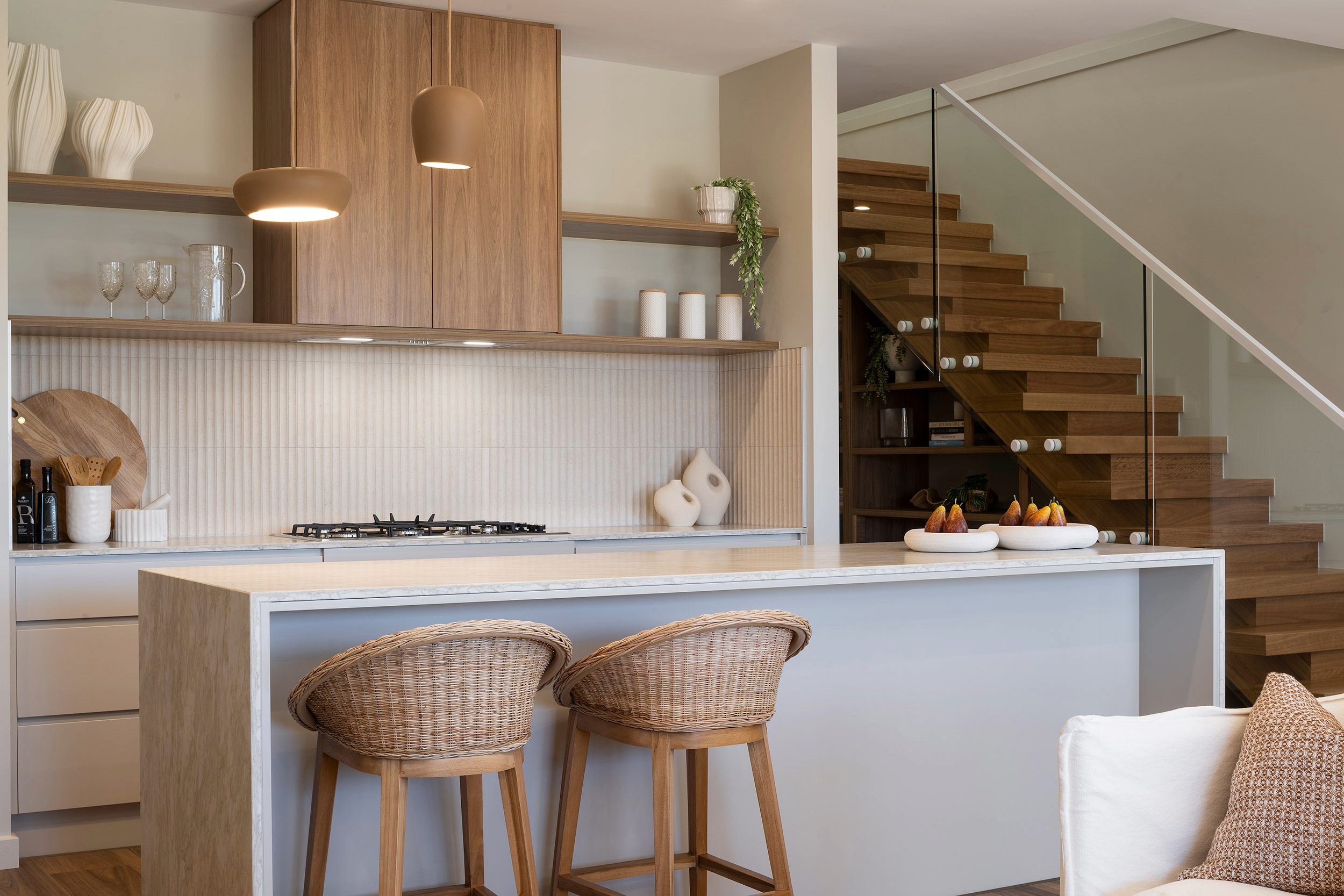 Modern luxury kitchen and living space with light blue island featuring two woven rattan chairs, gas cooktop, wooden pendant lights, open shelving with decorative items, and wooden staircase with glass railing visible in background.
