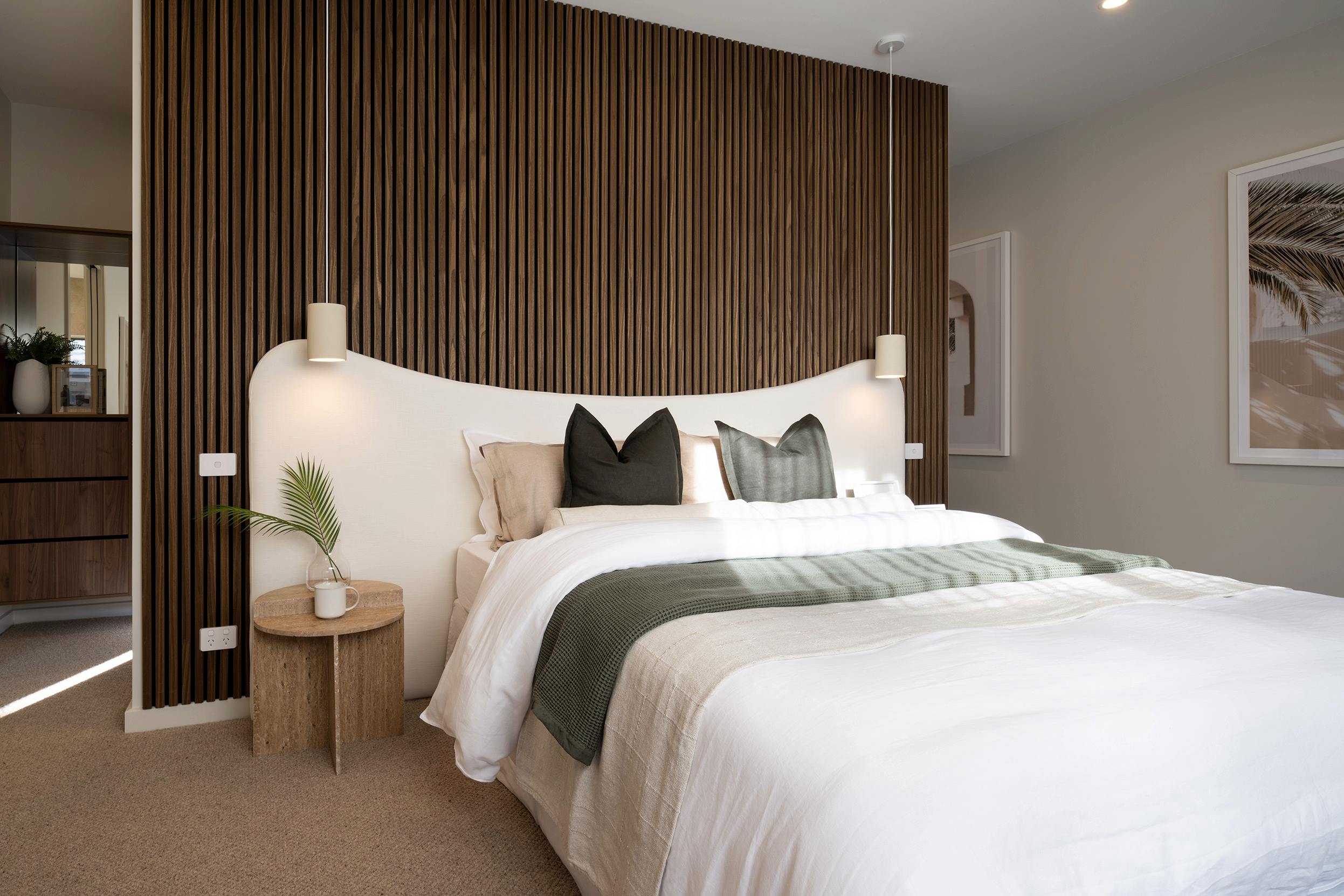 Modern luxury resort bedroom with a king bed featuring white linens, black and green decorative pillows, and a curved white headboard backed by vertical wood slat paneling. A wooden round side table with a potted plant sits beside the bed. The neutral color palette includes beige carpet, light gray walls, and warm wood furnishings. Pendant lights flank the headboard, and framed artwork hangs on the right wall.