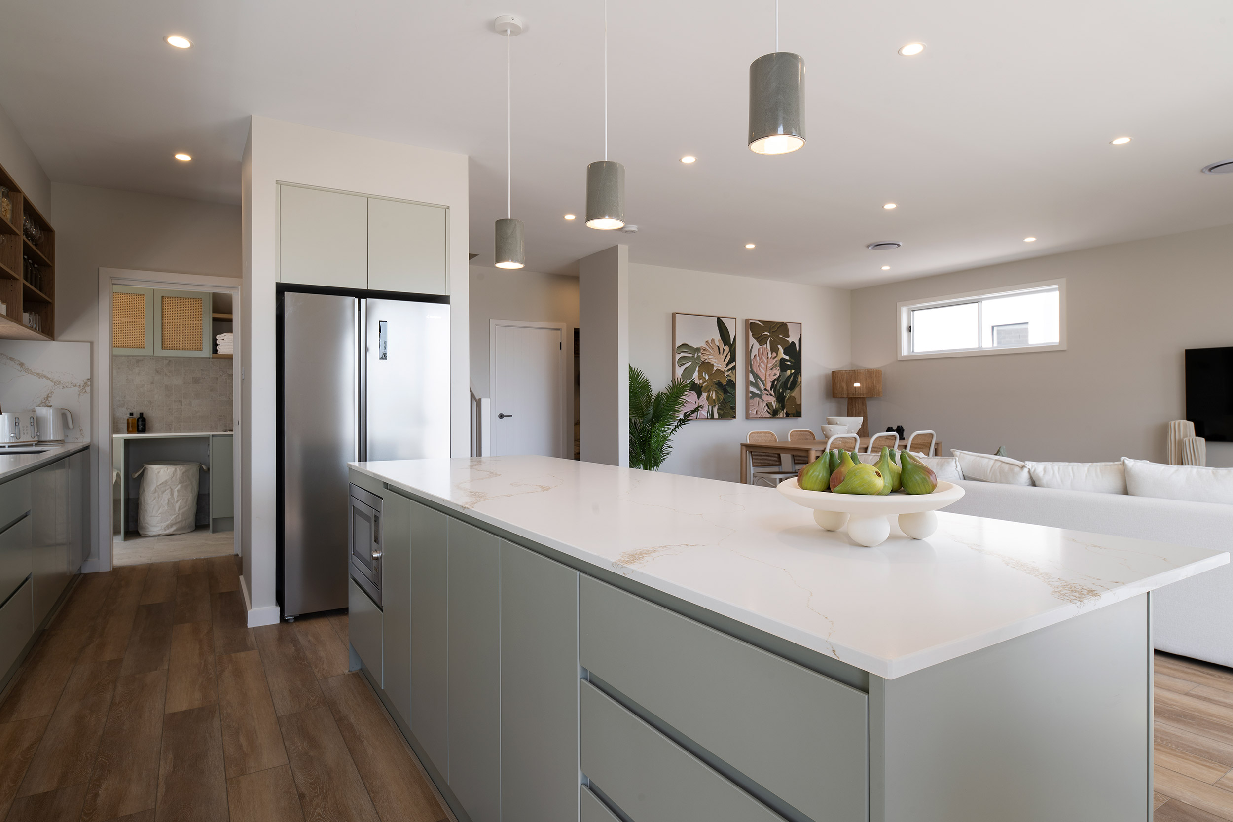 Modern luxury kitchen and open-plan living area featuring a large white marble island with green pears in a bowl, sage green cabinetry, stainless steel appliances, pendant lighting, and connected living spaces with botanical artwork and comfortable seating in the background.