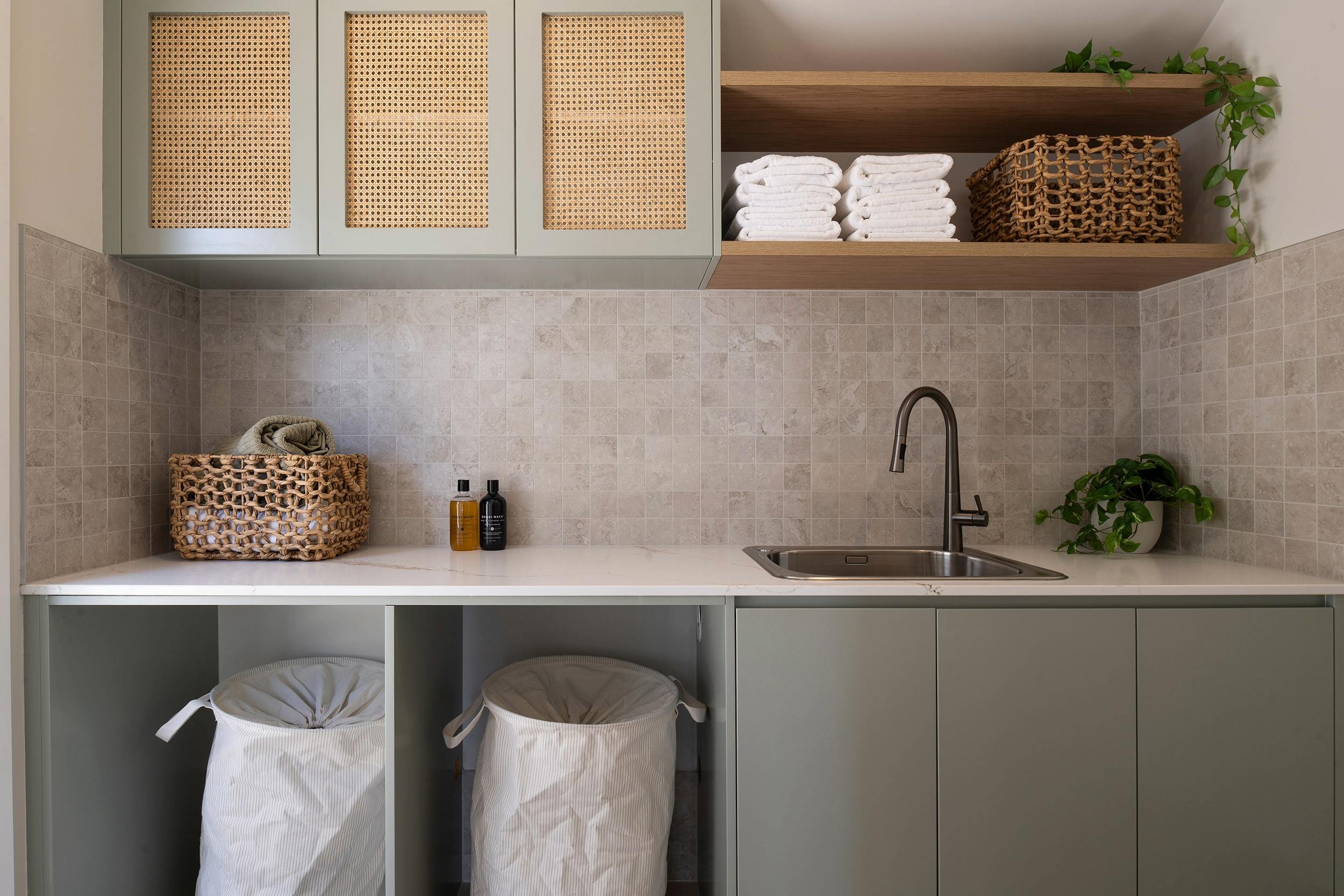 Modern luxury bathroom vanity with sage green cabinetry, white countertop, and black faucet. Upper cabinets feature woven rattan door panels and open shelving displaying folded white towels, woven baskets, and trailing green plants. Lower cabinets contain two fabric laundry hampers. Beige tile backsplash, stainless steel sink, and decorative bottles and potted plant on countertop.