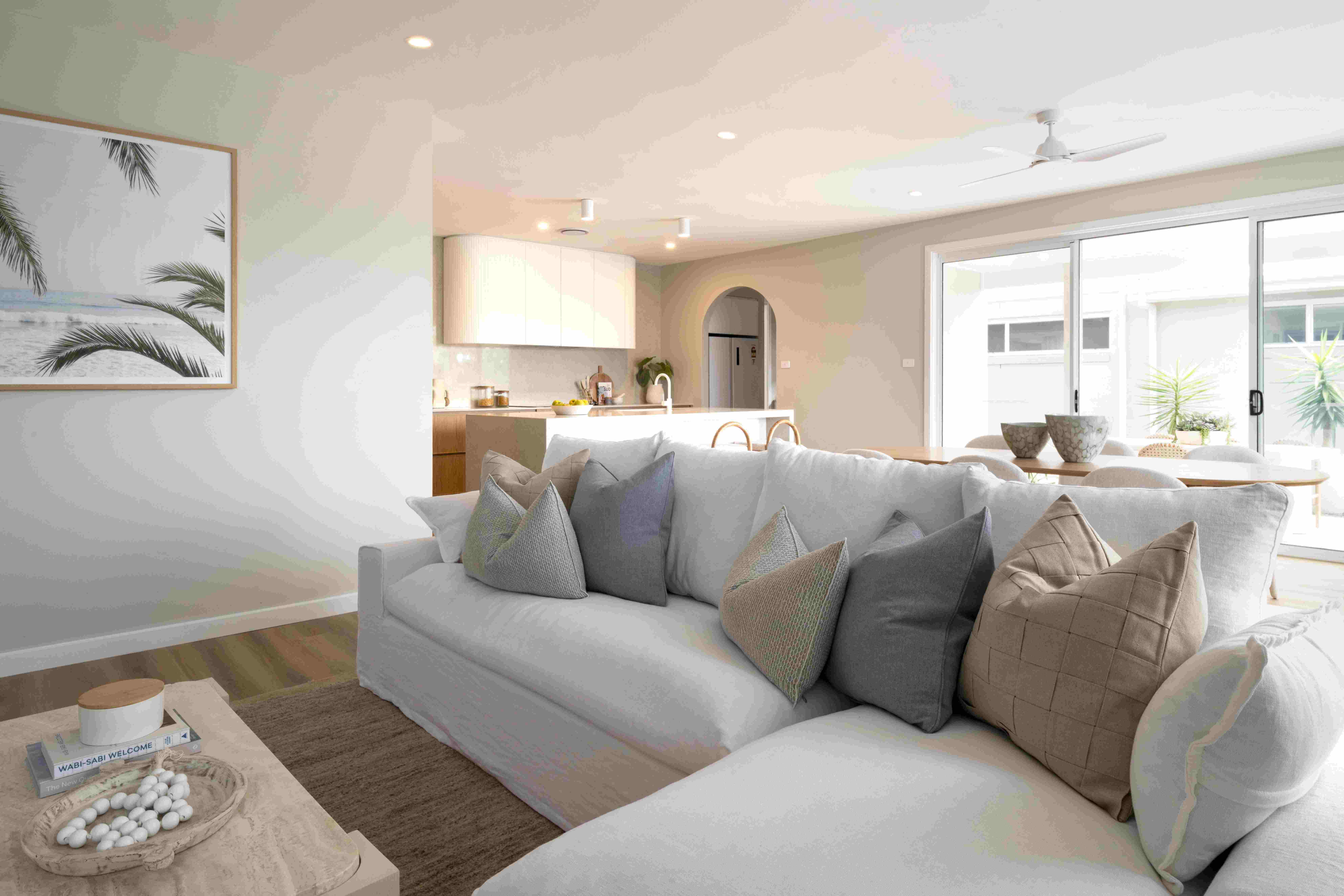 Modern luxury resort living room with a large light gray sectional sofa adorned with decorative pillows, facing a wooden coffee table with a woven bowl and welcome book. Behind the sofa is an open-plan kitchen with light wood cabinetry and white countertops. The space features floor-to-ceiling glass doors opening to an outdoor area with plants, neutral wall art with palm motifs, and contemporary ceiling fans. Natural light floods the bright, minimalist interior designed