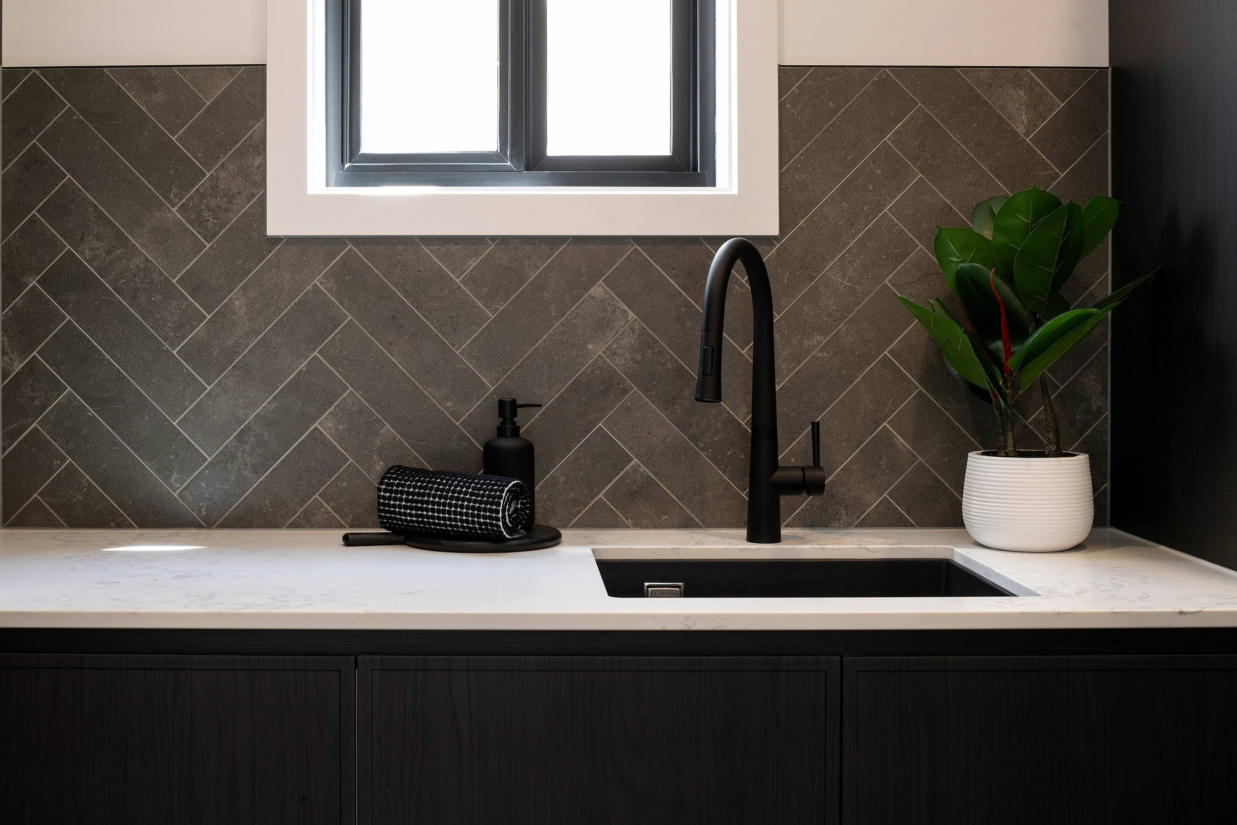 Modern bathroom vanity featuring a light marble countertop with a black undermount sink and matte black faucet. Dark herringbone-patterned tile backsplash extends to the wall behind. A window with white frame and dark trim sits above the counter. Black cabinetry with vertical paneling forms the base. A white textured pot with green plant and black soap dispenser with hand towel accessorize the counter.