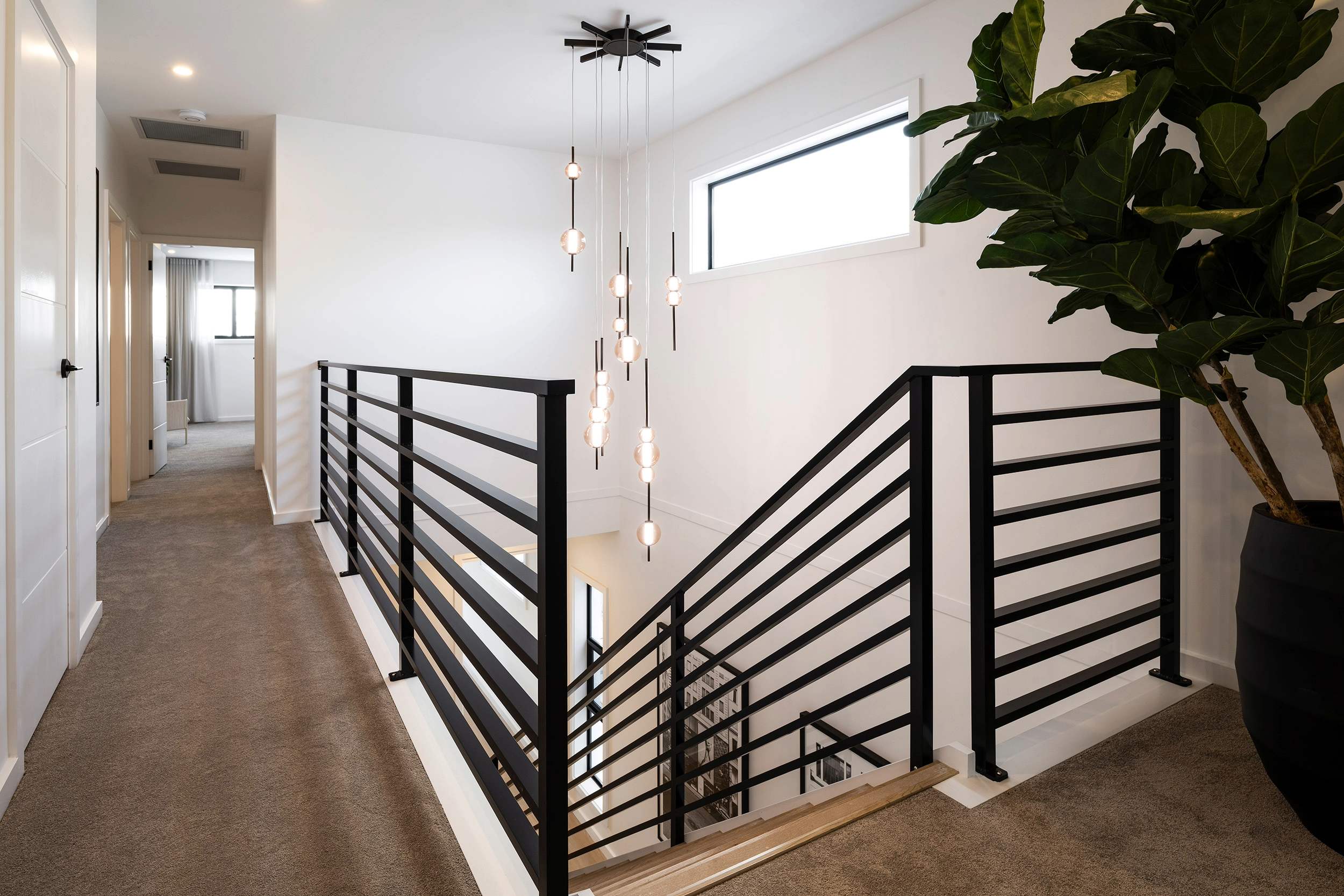 Modern hallway with black horizontal slat railings, pendant light fixture with cream-colored shades, white walls, large window, potted green plant on right side, and gray carpet flooring leading to white doors and bedroom area.