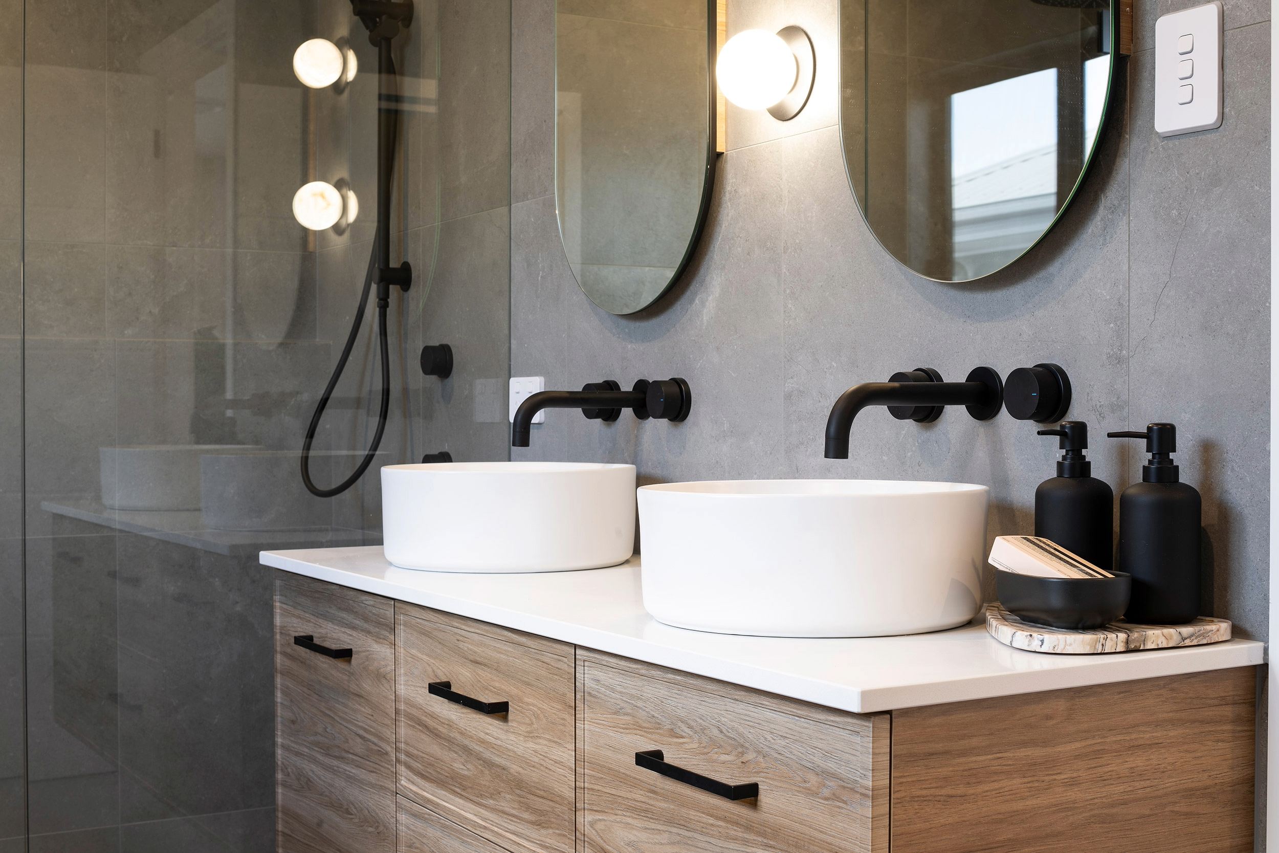 Modern bathroom vanity with dual white circular vessel sinks, black wall-mounted faucets, and illuminated mirrors. Wood-grain cabinetry with white countertop, black soap dispensers, and decorative accessories. Glass shower enclosure visible on left with black fixtures and rainfall showerhead.