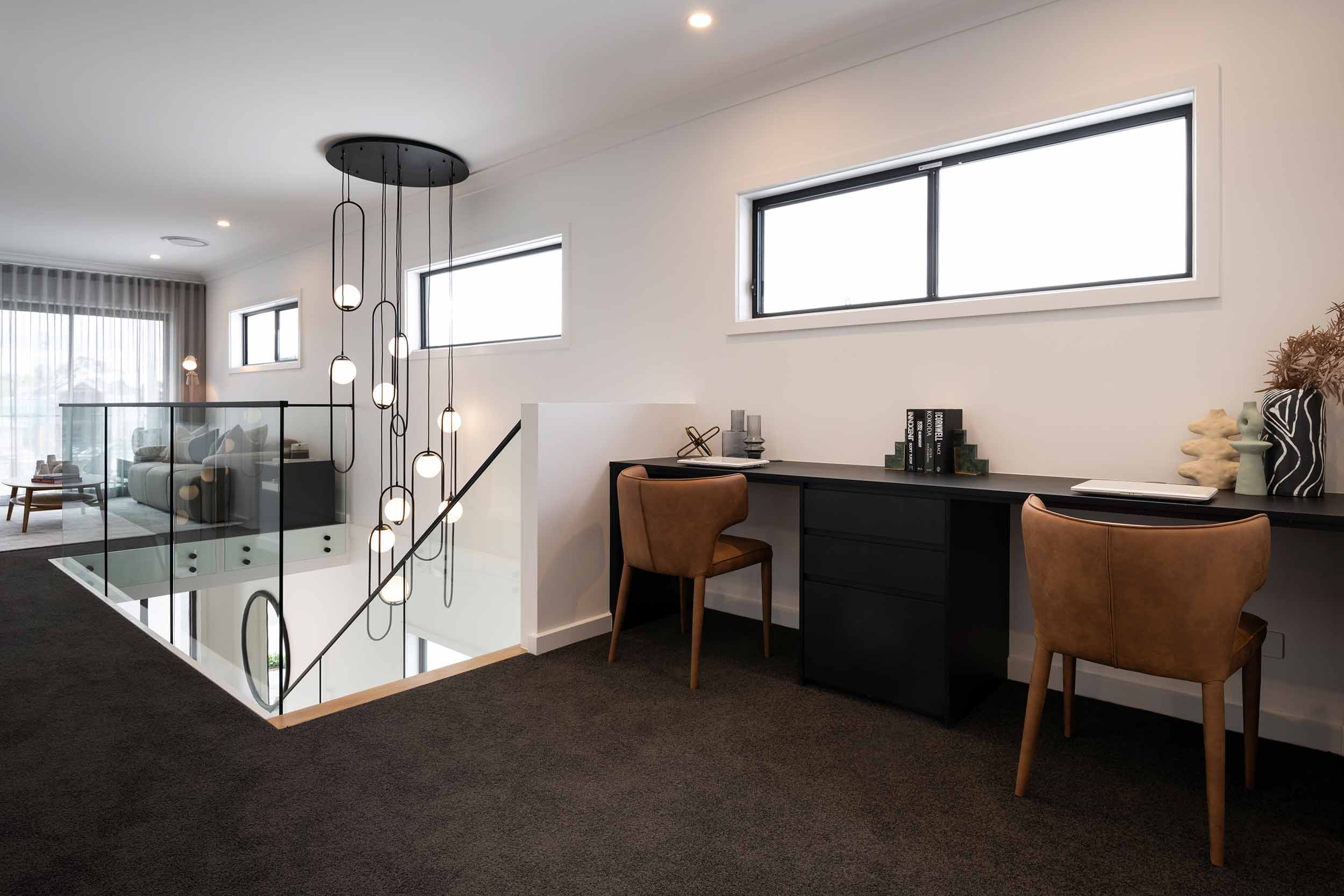 Modern home office with black desk, two cognac leather chairs, and contemporary pendant lighting. Large windows with black frames provide natural light. Glass railing with black metal handrail overlooks lower level. Neutral walls, dark carpet, and decorative accessories including books and plants complete the elegant workspace.