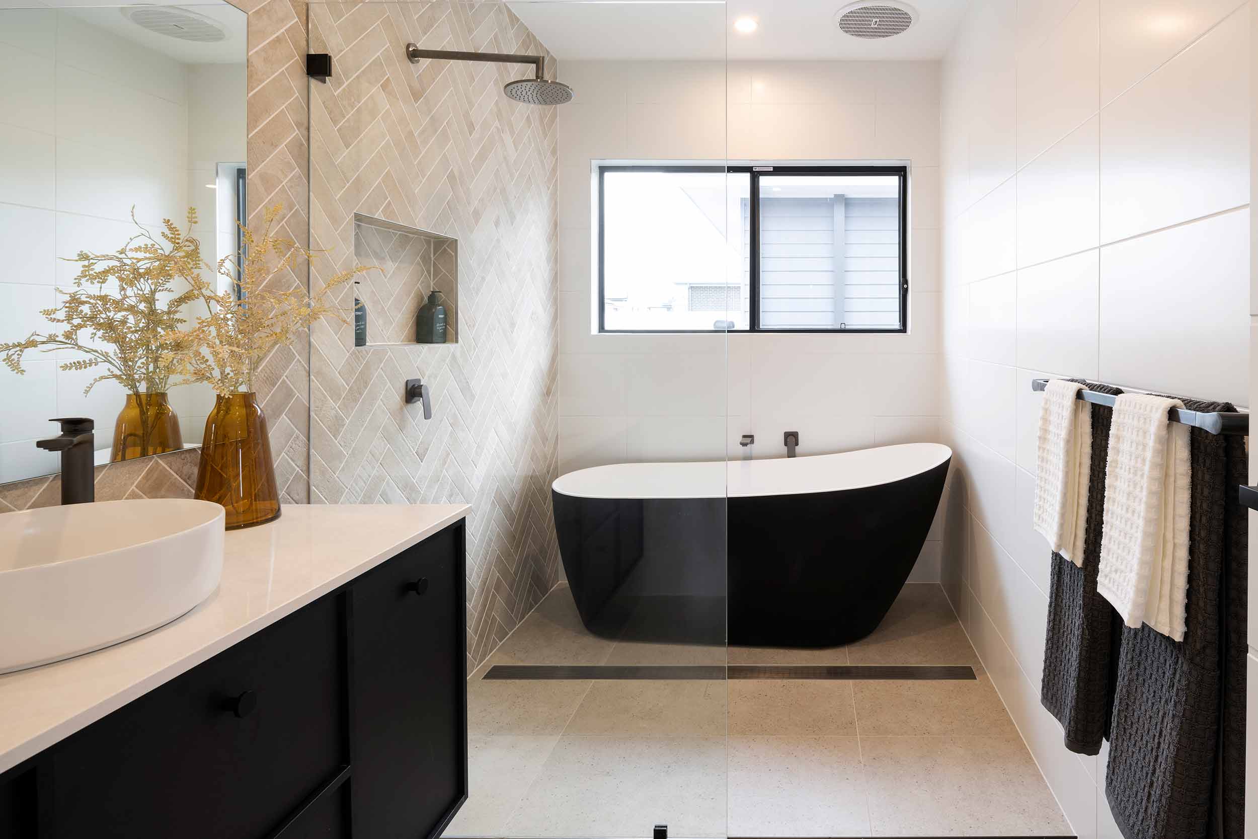 Modern bathroom with black and white color scheme featuring a freestanding black soaker tub with white interior, herringbone pattern tile shower wall, floating black vanity with white vessel sink, large window with black frame, rainfall showerhead, towel rack with folded towels, and decorative amber glass vases with dried botanicals.