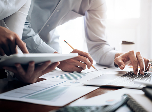 Two professionals reviewing tender documents together at a desk, with one person pointing at a spreadsheet while the other uses a laptop, surrounded by papers and a pen.